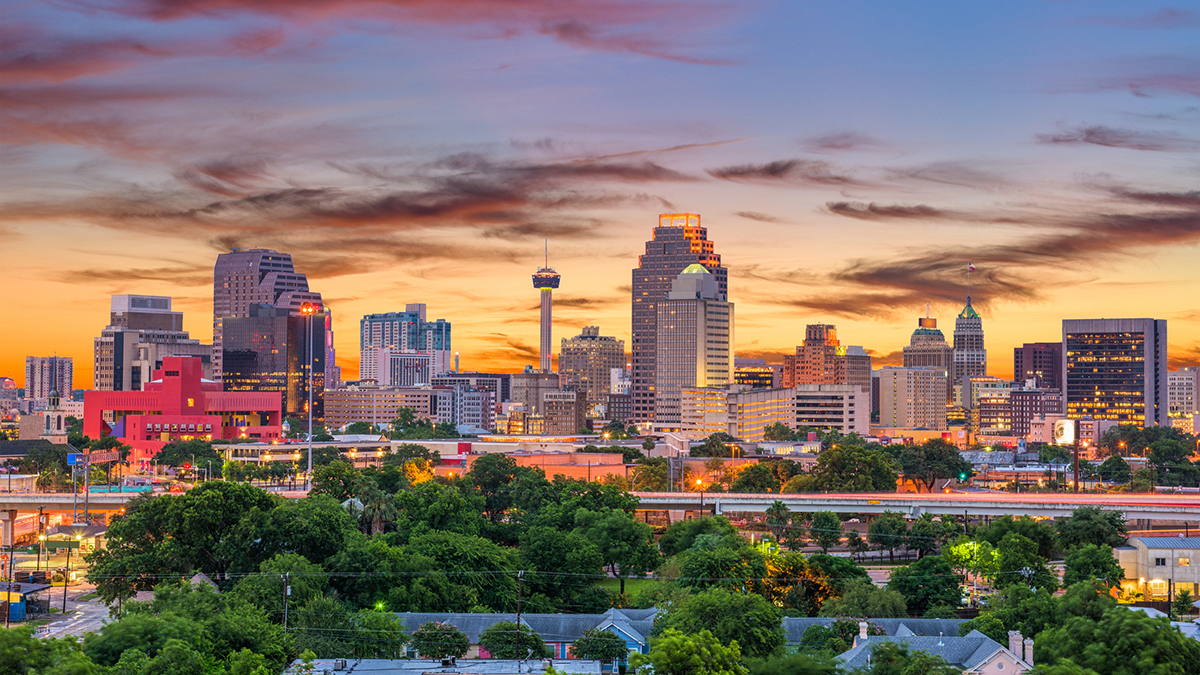 San Antonio, Texas skyline with some trees up front, then downtown with office buildings and in the back a sunset sky with a few clouds.