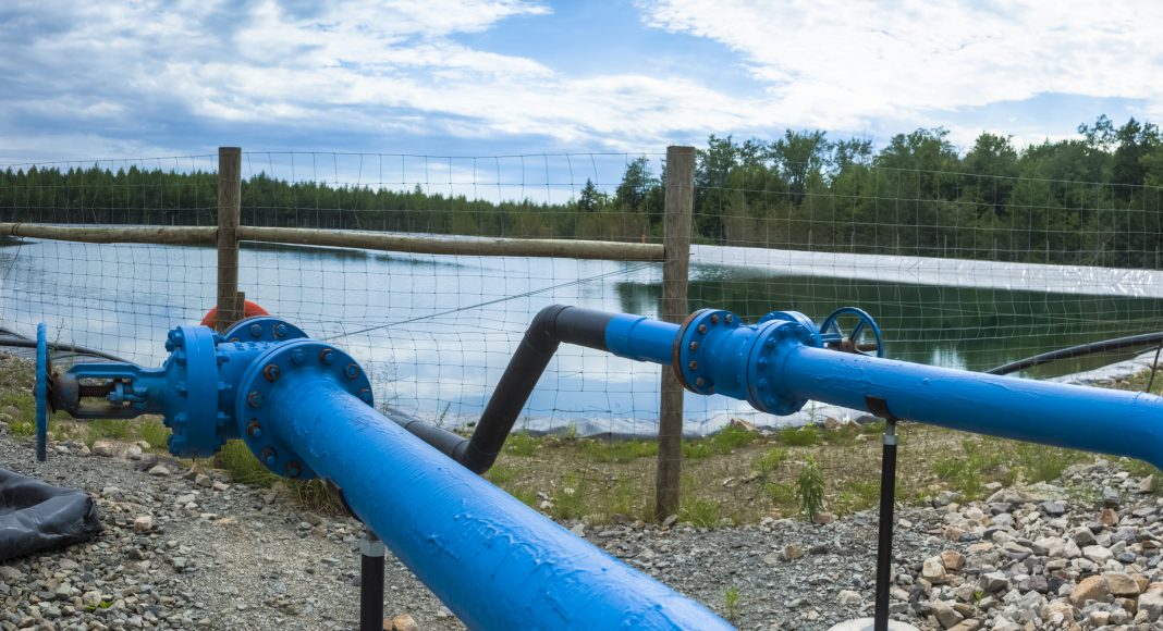 A man-made pond with pine trees in the background. Blue pipes leading to the pond in the foreground.